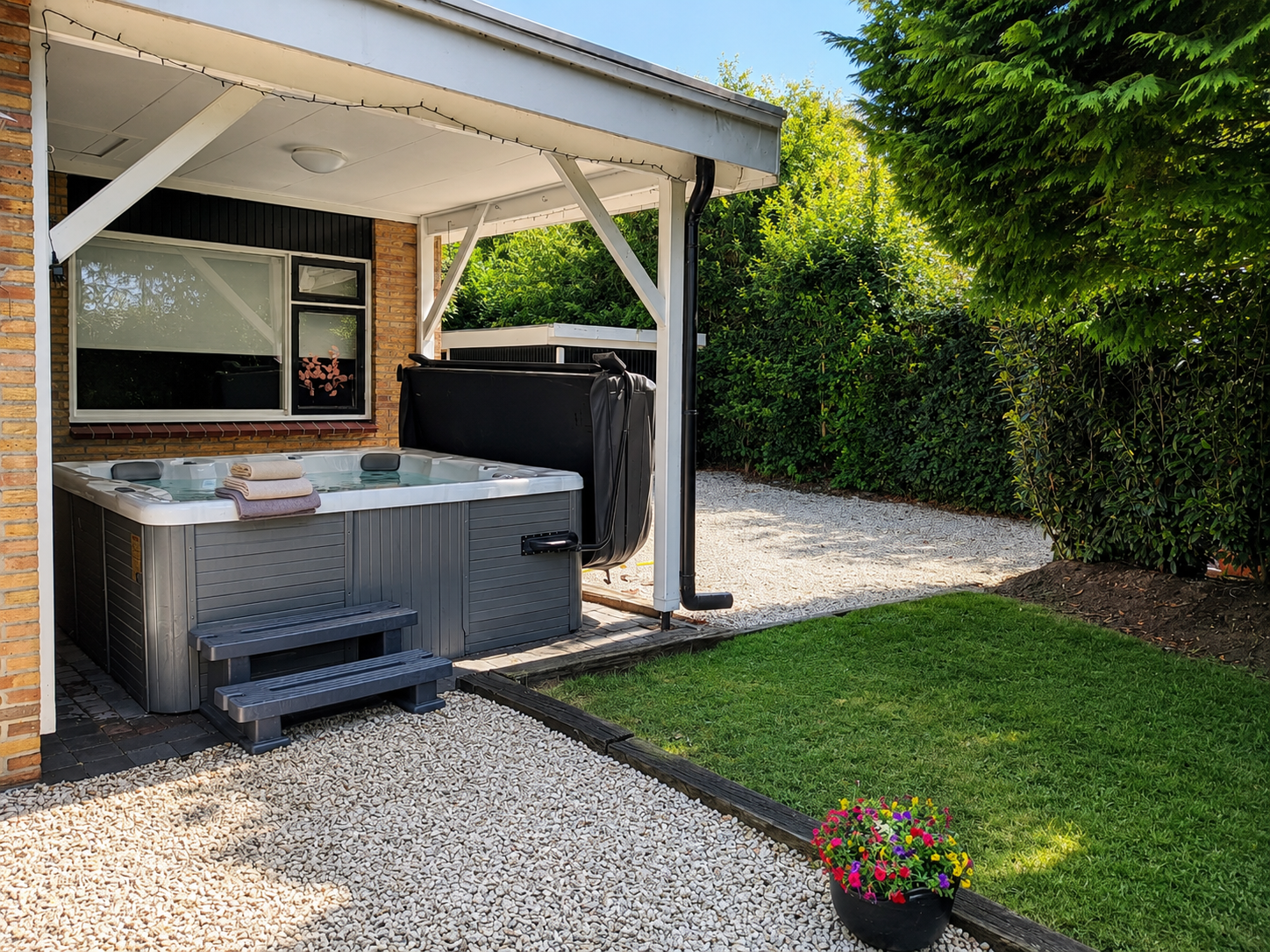 Overdekt terras met een jacuzzi, een kleine trap, een grindpad en een groen gazon naast een heg.