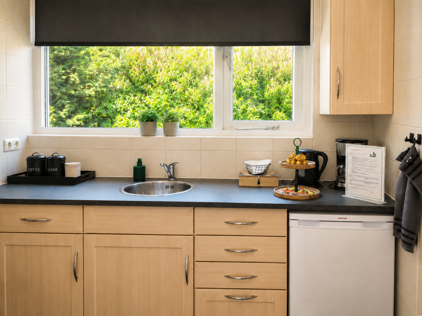 Lichte keuken met lichte houten kasten, een spoelbak bij een zonnig raam en zwarte aanrechtbladen.