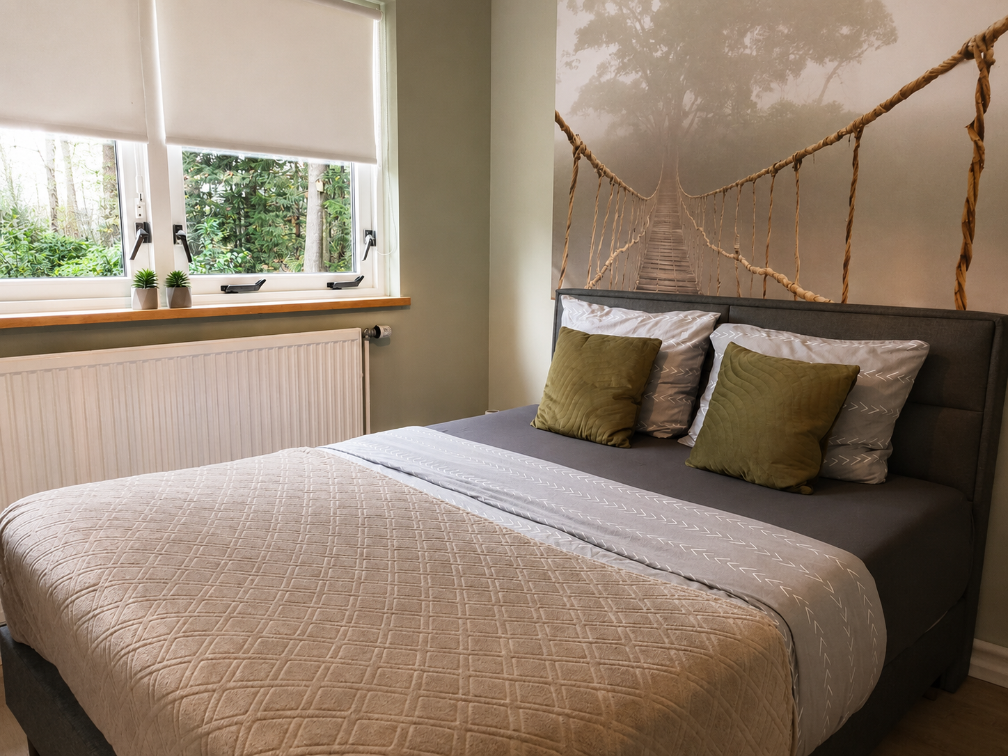 Gezellige slaapkamer met een bed, grijs beddengoed, groene kussens en een raam met planten en jaloezieën.