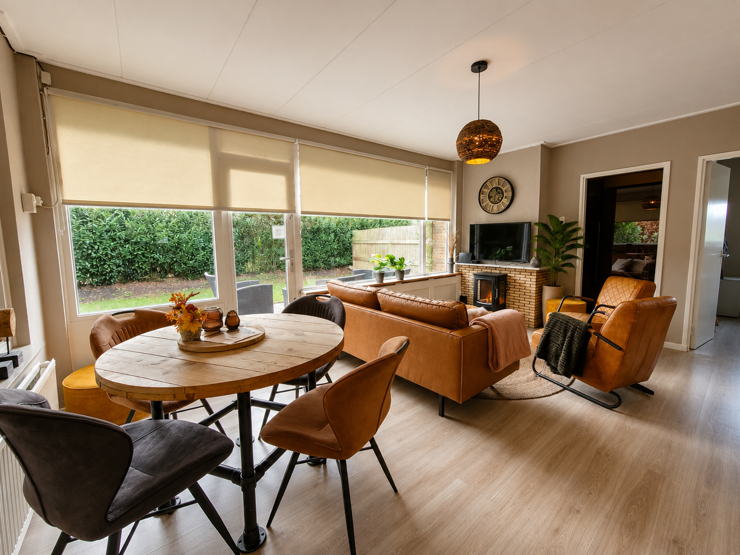 Lichte, moderne woonkamer met beige banken, ronde eettafel, grote ramen en een hanglamp met patroon.