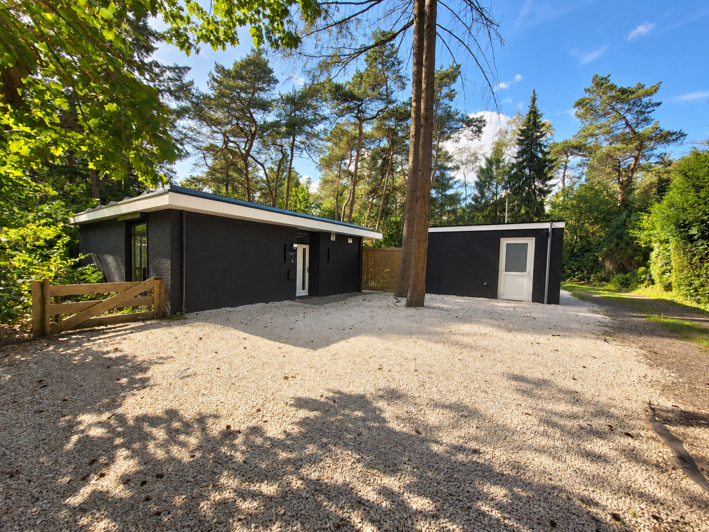 Moderne, donkergrijs huis in een open plek in het bos met een grindondergrond en hoge bomen.