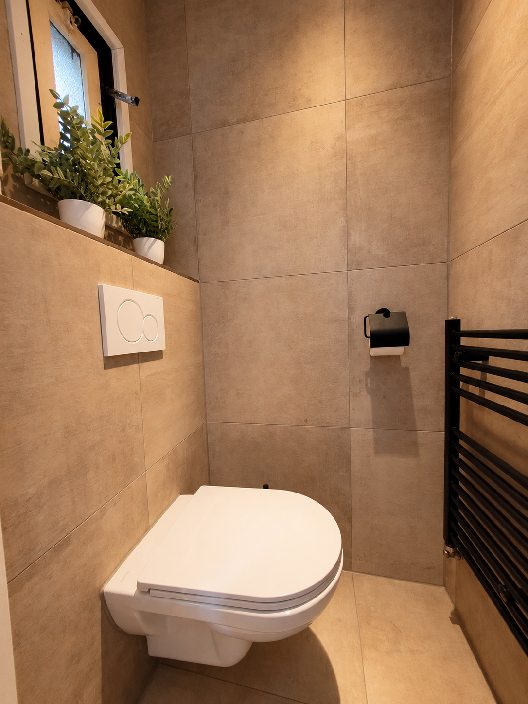 Moderne badkamer met een aan de muur gemonteerd wit toilet, beige betegelde muren en een klein raam met planten.