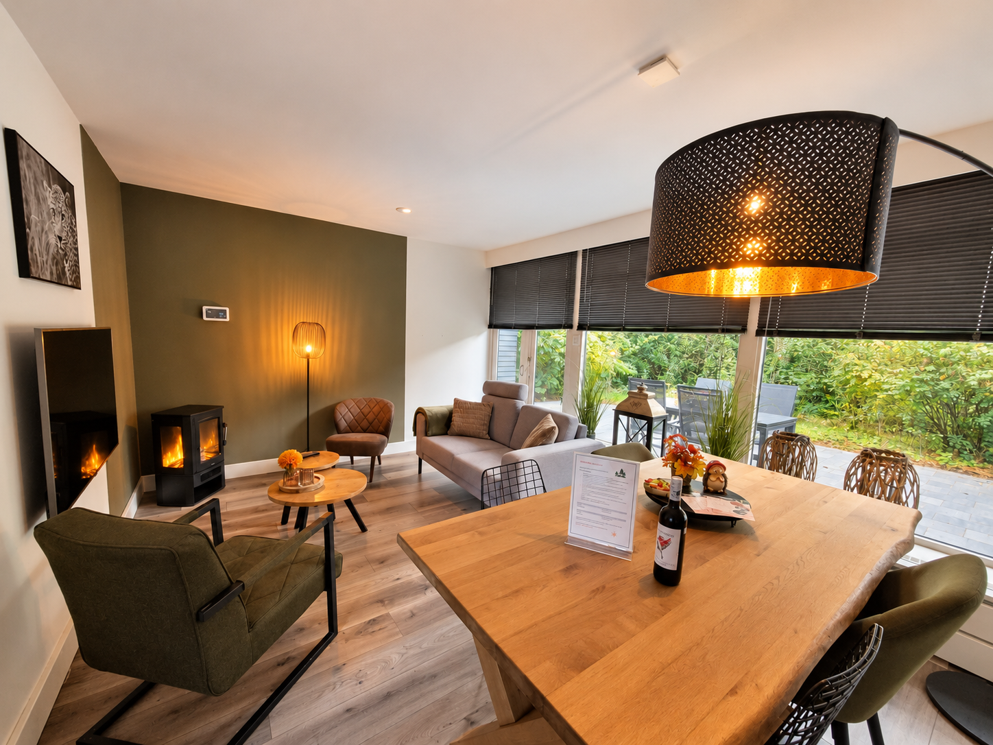 Moderne woonkamer met open haard, eettafel en grote ramen met uitzicht op het groen.