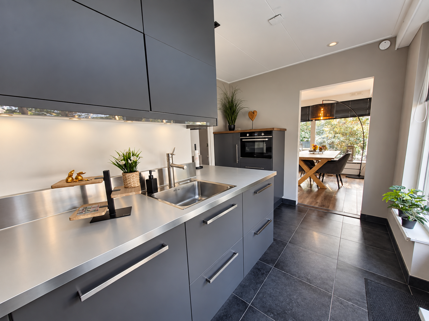 Moderne keuken met grijze kasten, roestvrijstalen aanrechtbladen en uitzicht op een lichte eetruimte.