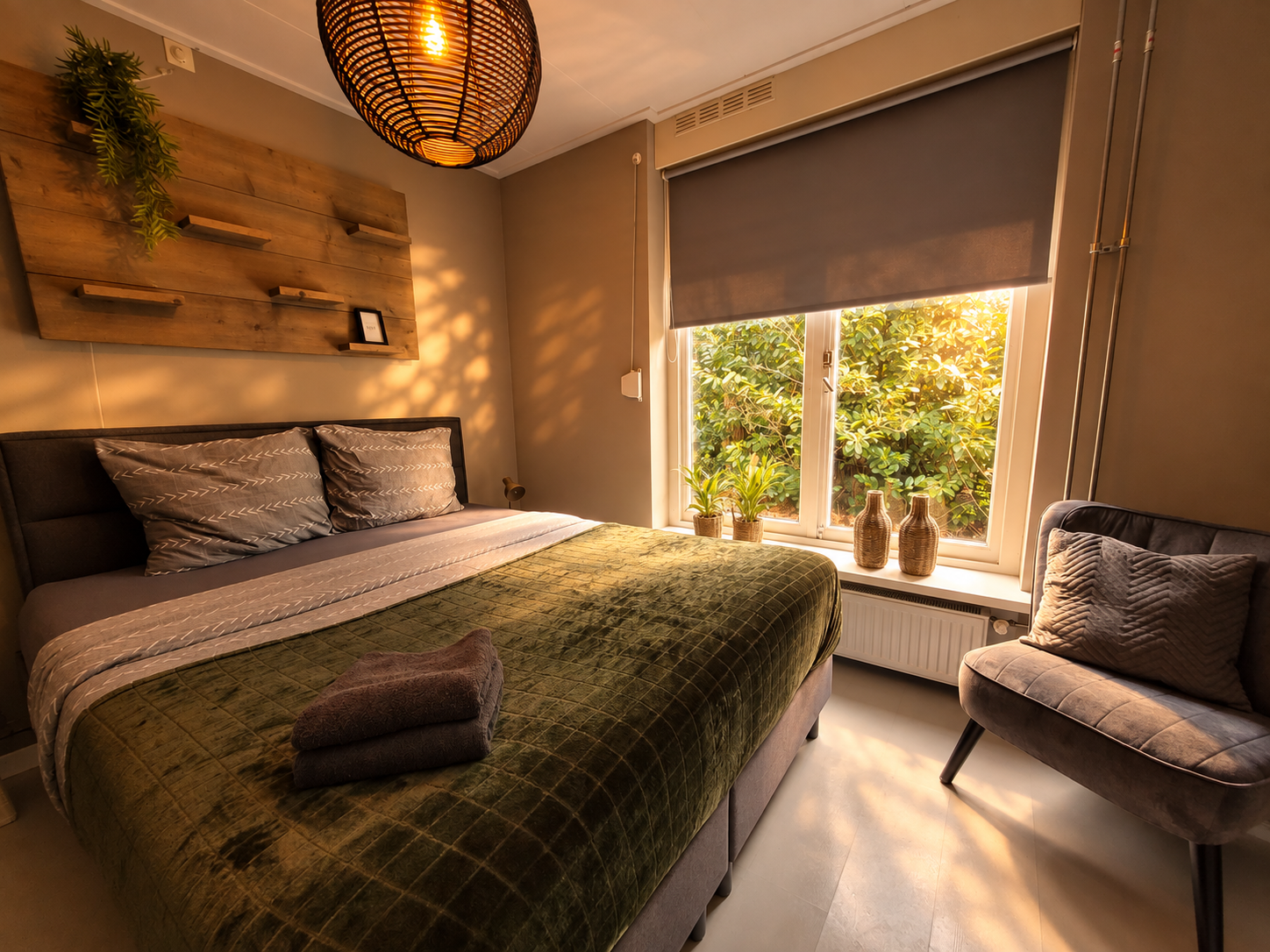 Gezellige slaapkamer met een bed, fauteuil, warme verlichting en een raam met een grijs rolgordijn waar de zon op schijnt.