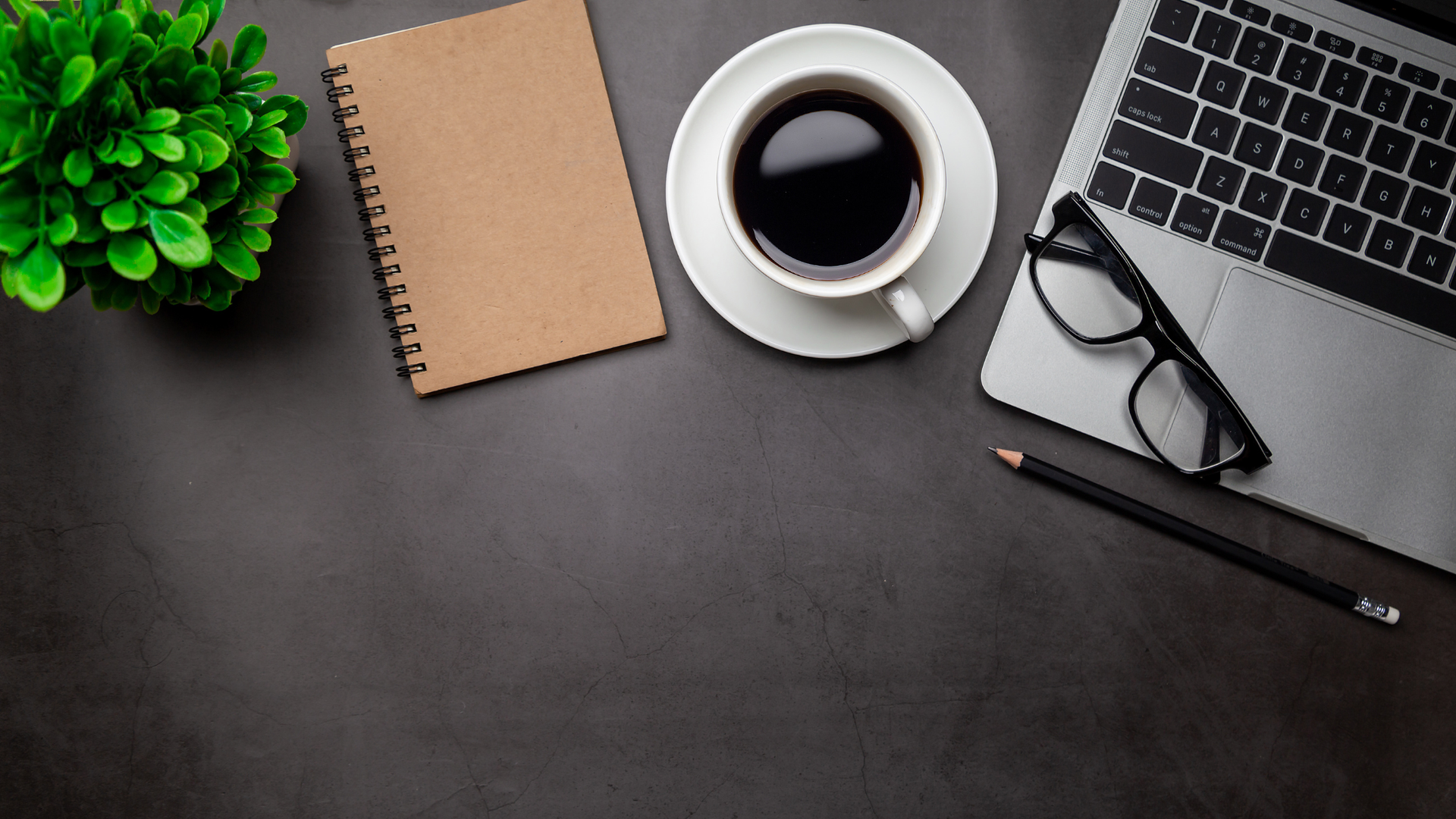 Overhead view of desk with coffee, laptop, glasses, notebook, plant, and pencil.