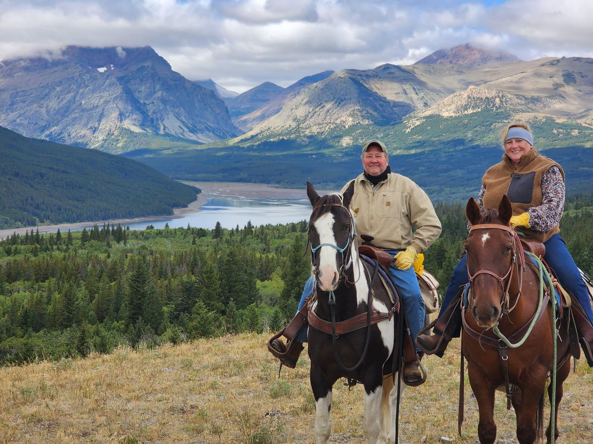Two men are riding horses in a field with mountains in the background.