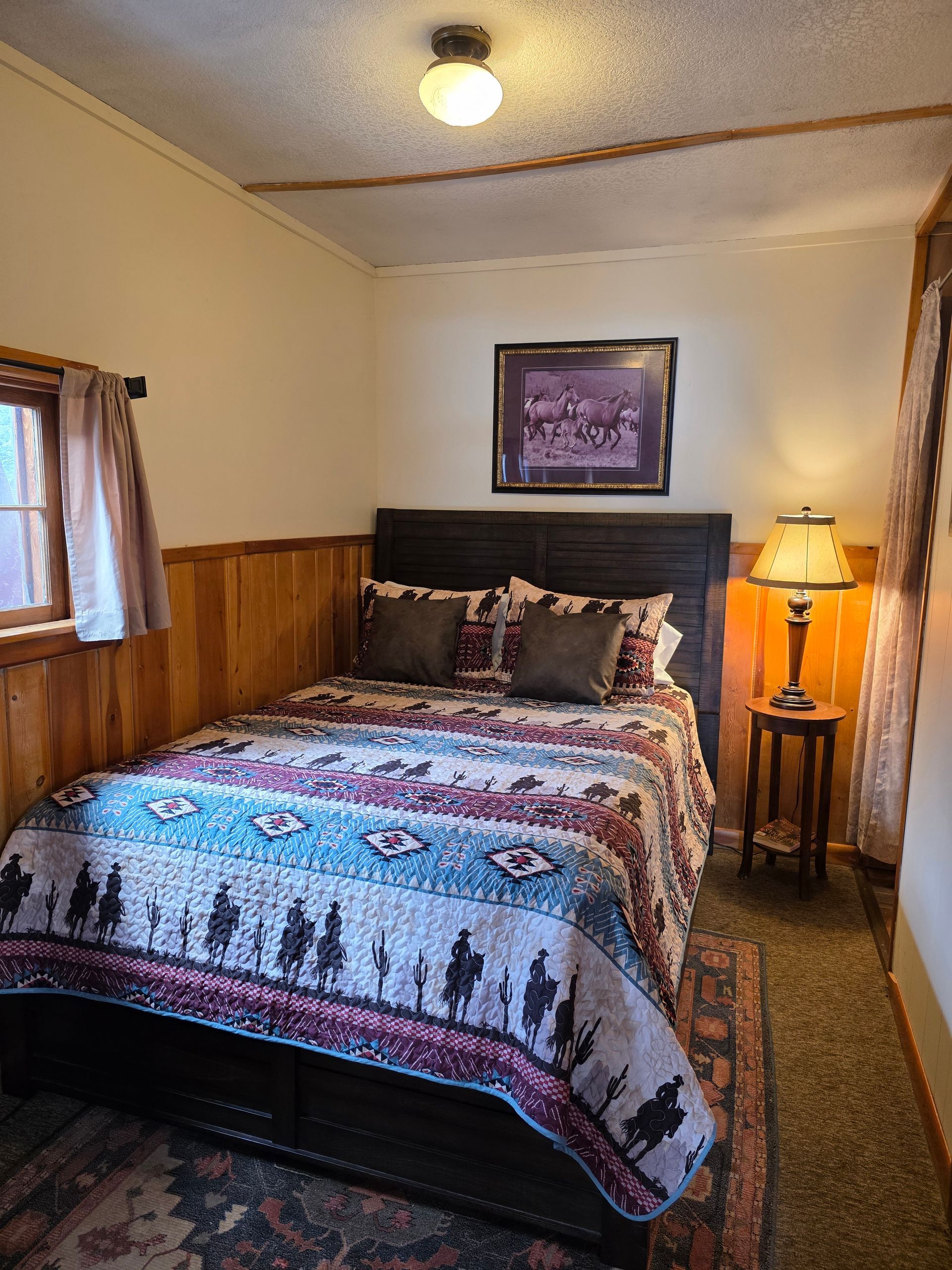 Bedroom with a bed, nightstand, and lamp. Western-themed bedspread and artwork. Wooden paneling on walls.