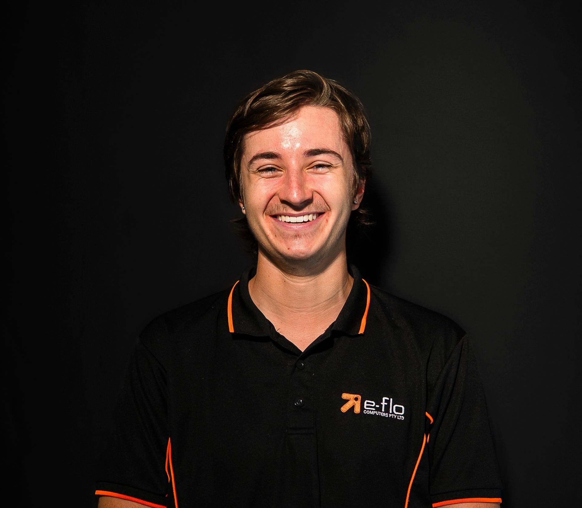 Smiling person in a black polo shirt with orange trim against a dark background
