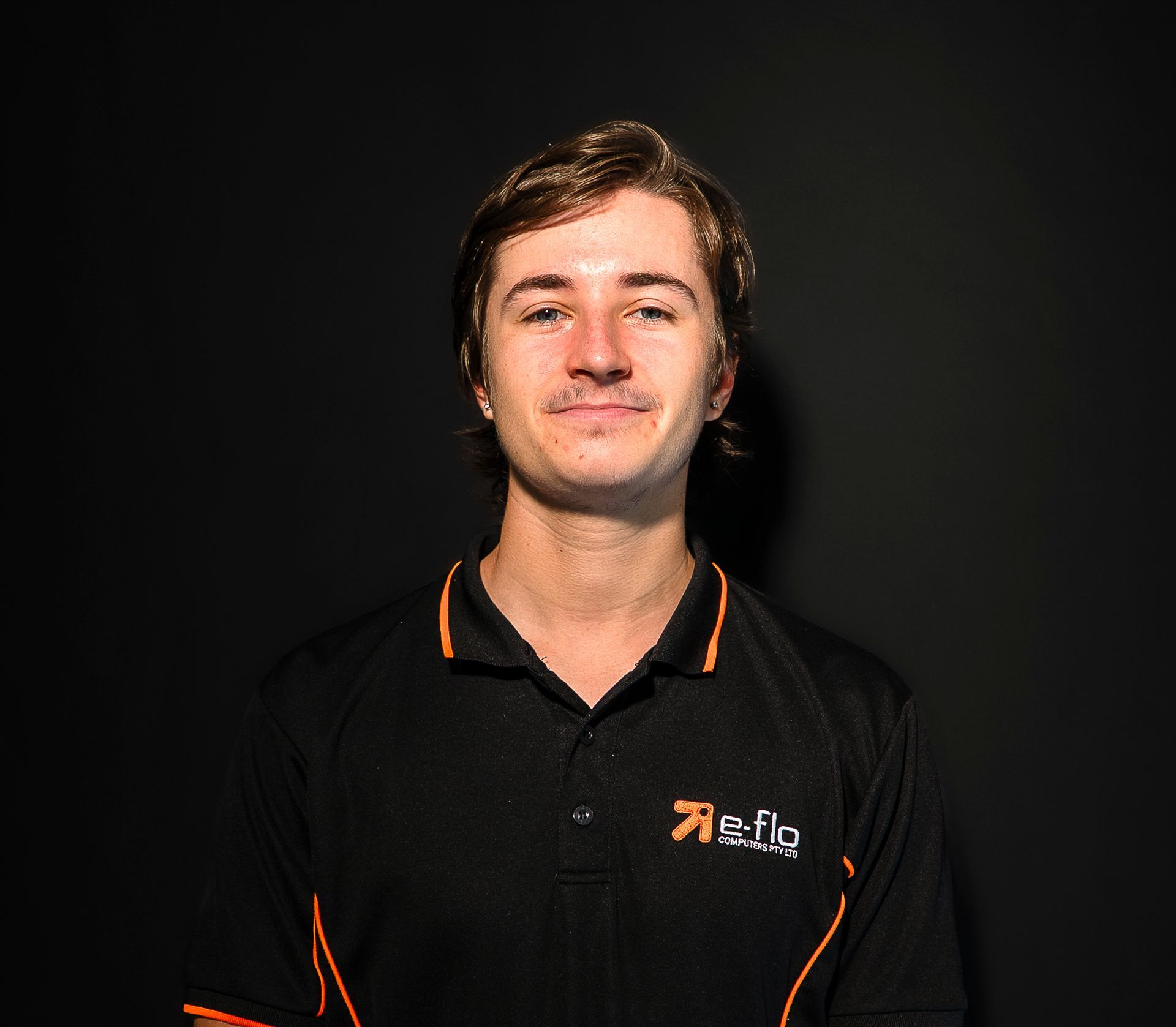 Man in a black polo shirt with orange trim, standing against a dark background and looking at the camera
