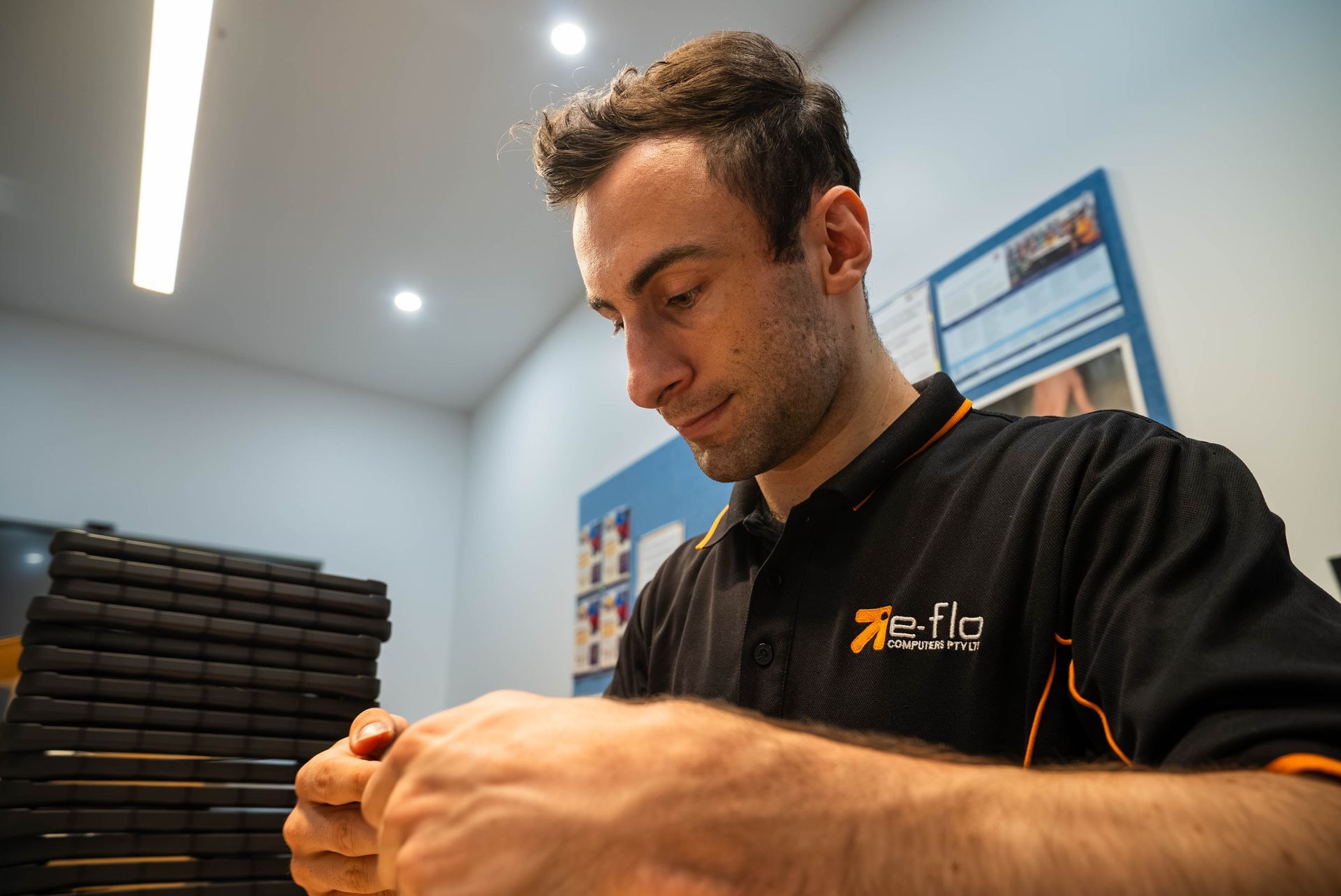 Man in a black work shirt handling tools in a bright workshop, with stacked items in the background