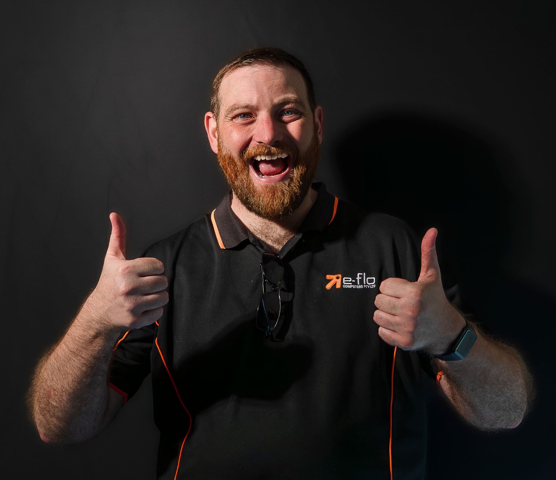 Smiling man with beard giving two thumbs up against a dark background, wearing a black shirt with red accents