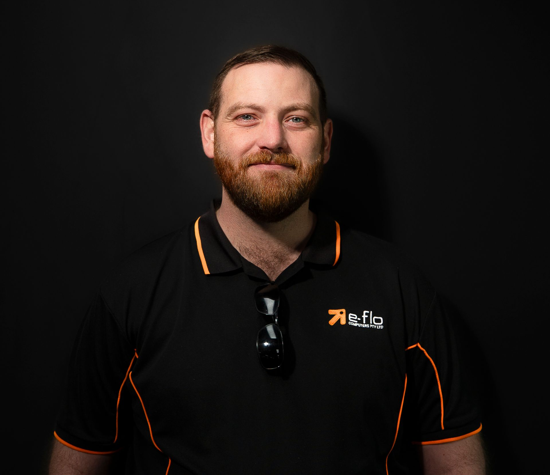 Man in black polo shirt with orange trim, standing against a dark background and smiling