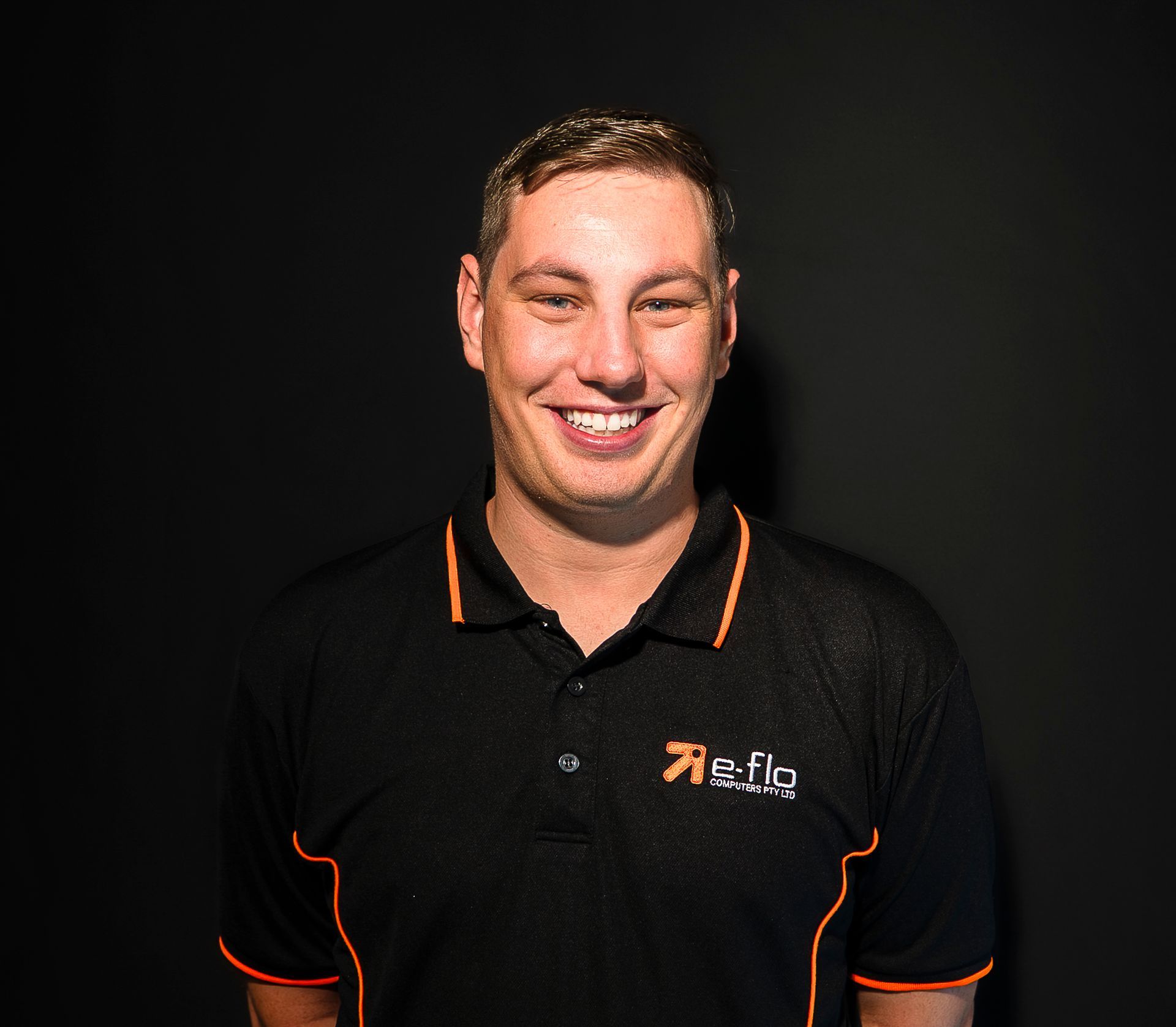 Smiling man in black polo shirt with orange trim against a dark background