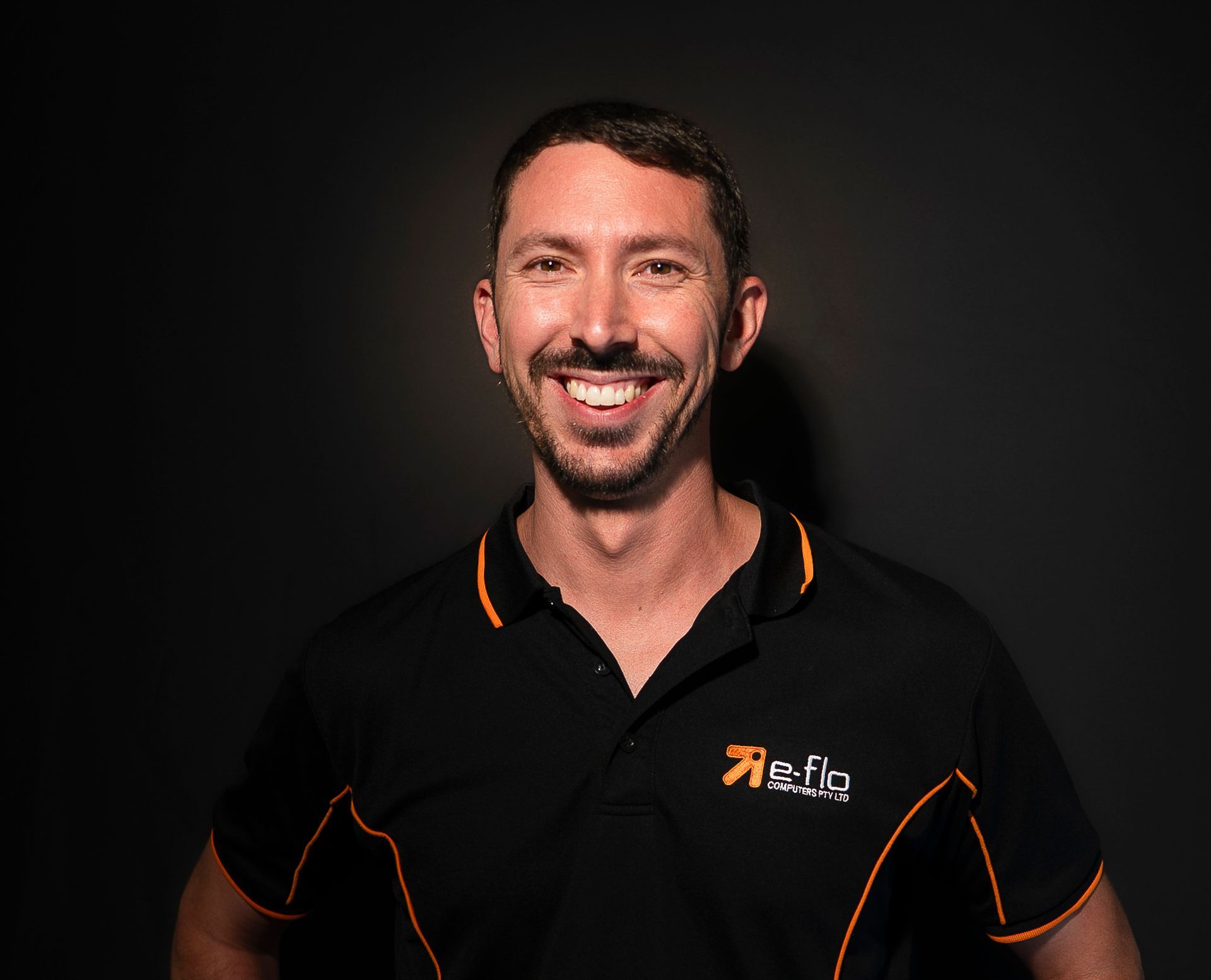 Smiling person in a black polo shirt with orange trim against a dark background