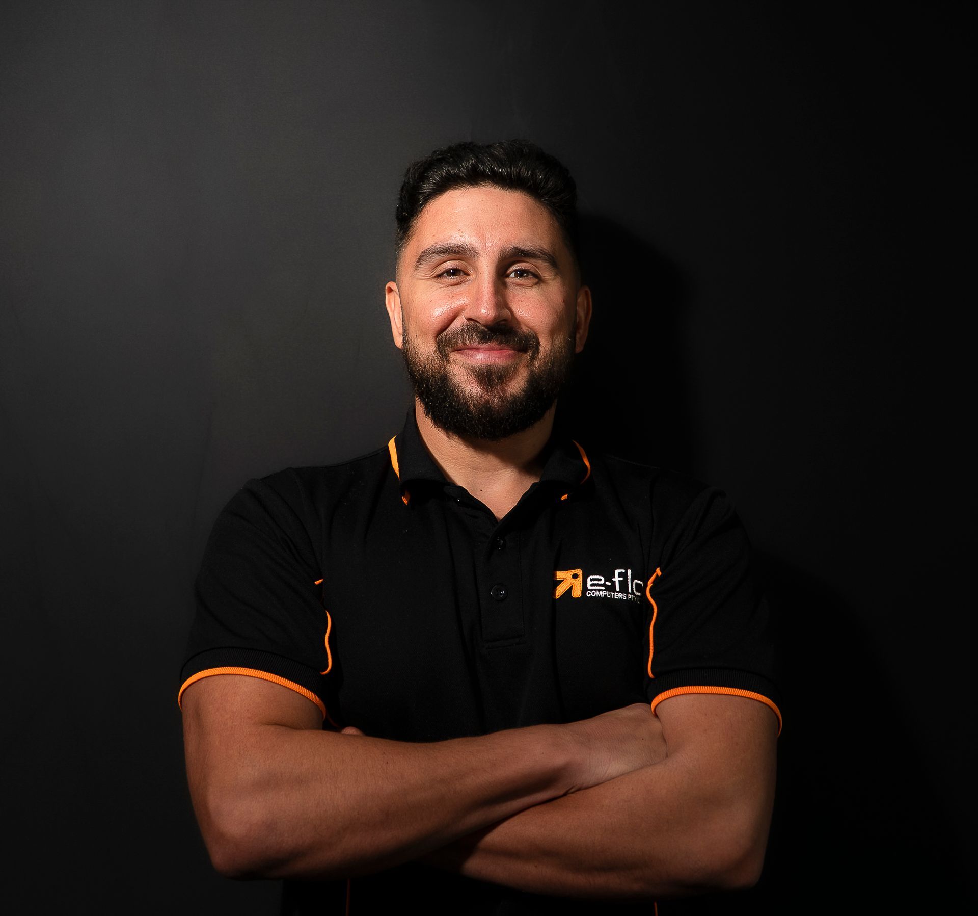 Portrait of a smiling man in a black polo with orange trim against a dark background