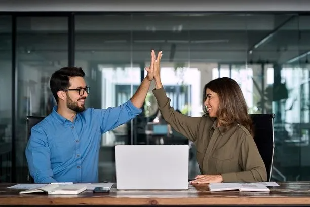 Two coworkers smiling and discussing a laptop at a table in a modern office