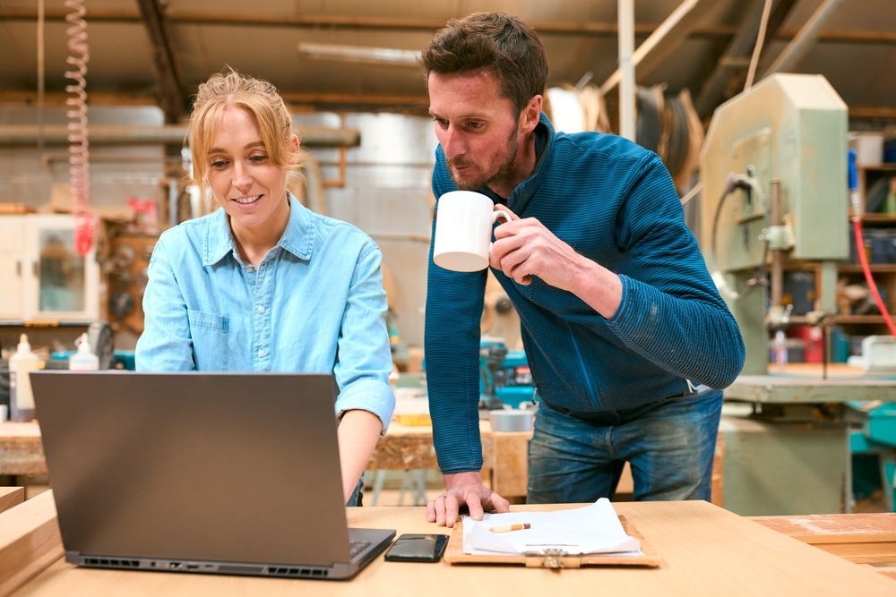Two coworkers review a laptop at a workbench in a workshop, one holding a mug and leaning in.