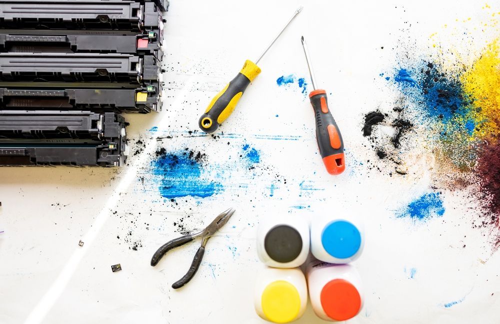 Printer toner cartridges and repair tools with spilled colorful ink on a white surface