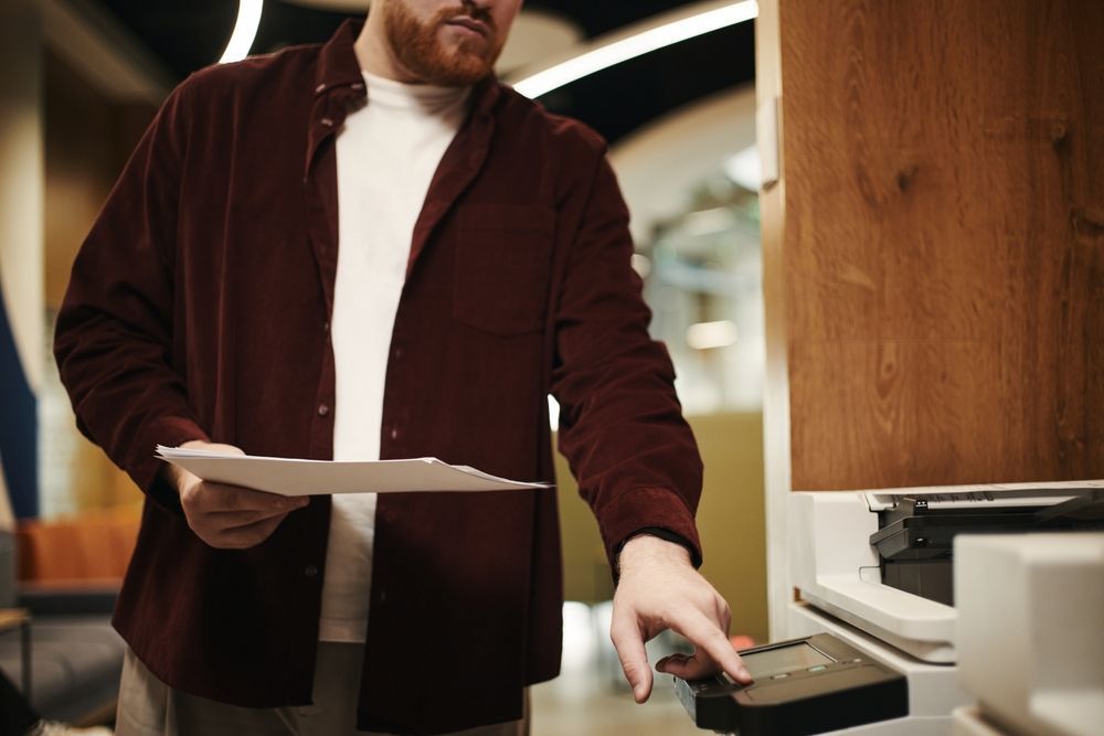 Person in a maroon jacket holding papers and pressing a button at a kiosk or printer.
