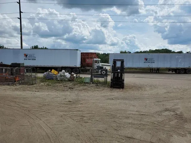 Two semi-trailers parked on a dirt lot with a forklift and debris in the foreground under a cloudy sky.