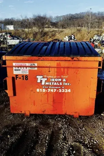 A bright orange commercial dumpster labeled TT Iron & Metals Inc. sitting on a dirt and gravel lot.