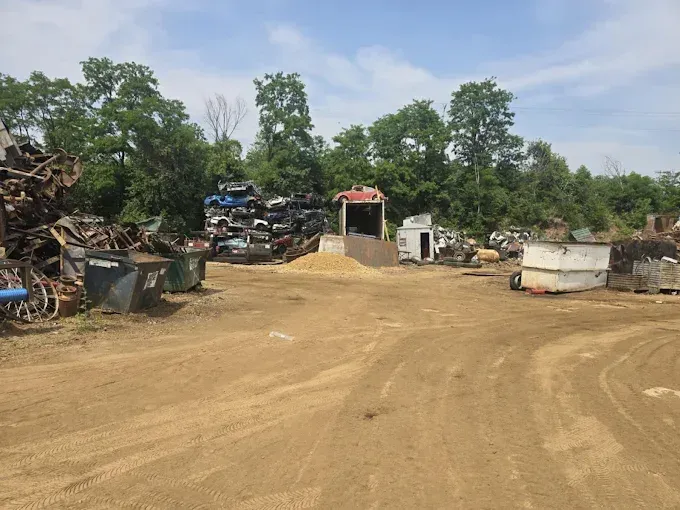 A scrapyard with piles of discarded metal and car parts on a dirt lot, framed by trees under a blue sky.