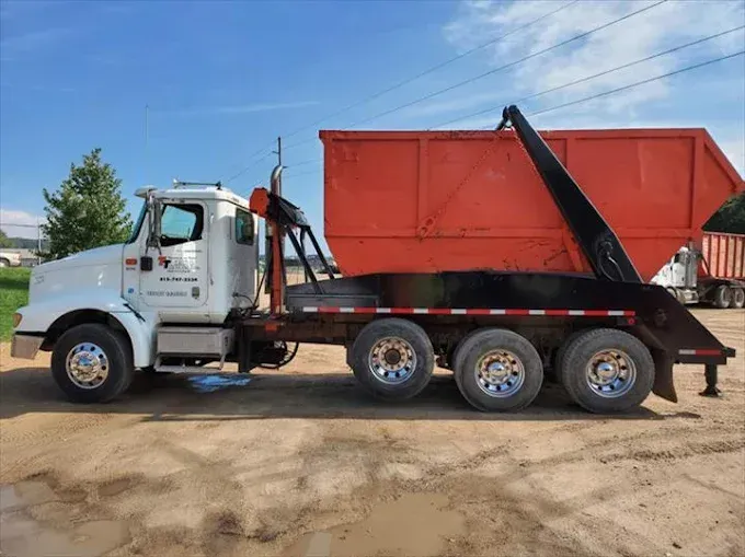 A white roll-off truck carrying a large orange dumpster on a dirt lot under a blue sky.
