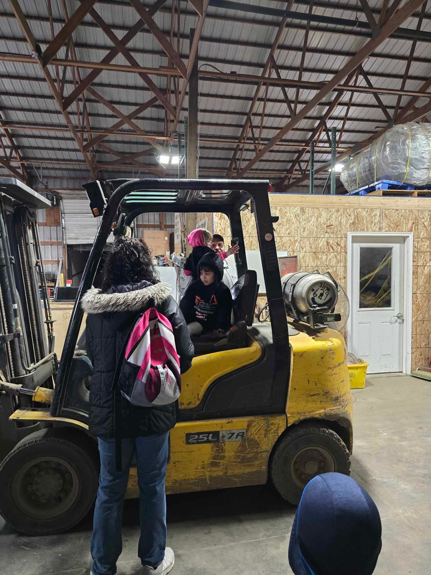 A person with a pink backpack stands next to a child sitting in the driver’s seat of a yellow forklift in a warehouse.