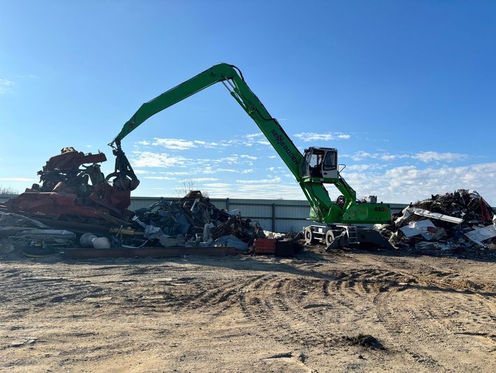 A bright green scrap material handler with a grapple arm moves metal debris at a recycling yard under a clear blue sky.