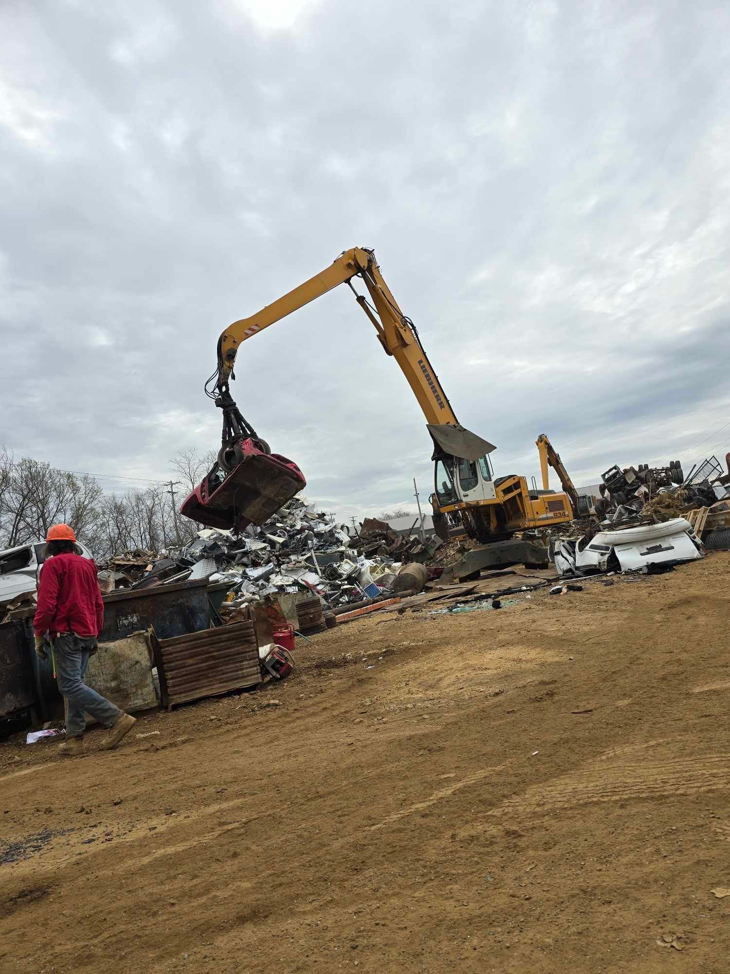 A yellow excavator with a grapple attachment lifts a piece of scrap metal in a recycling yard under a cloudy sky.