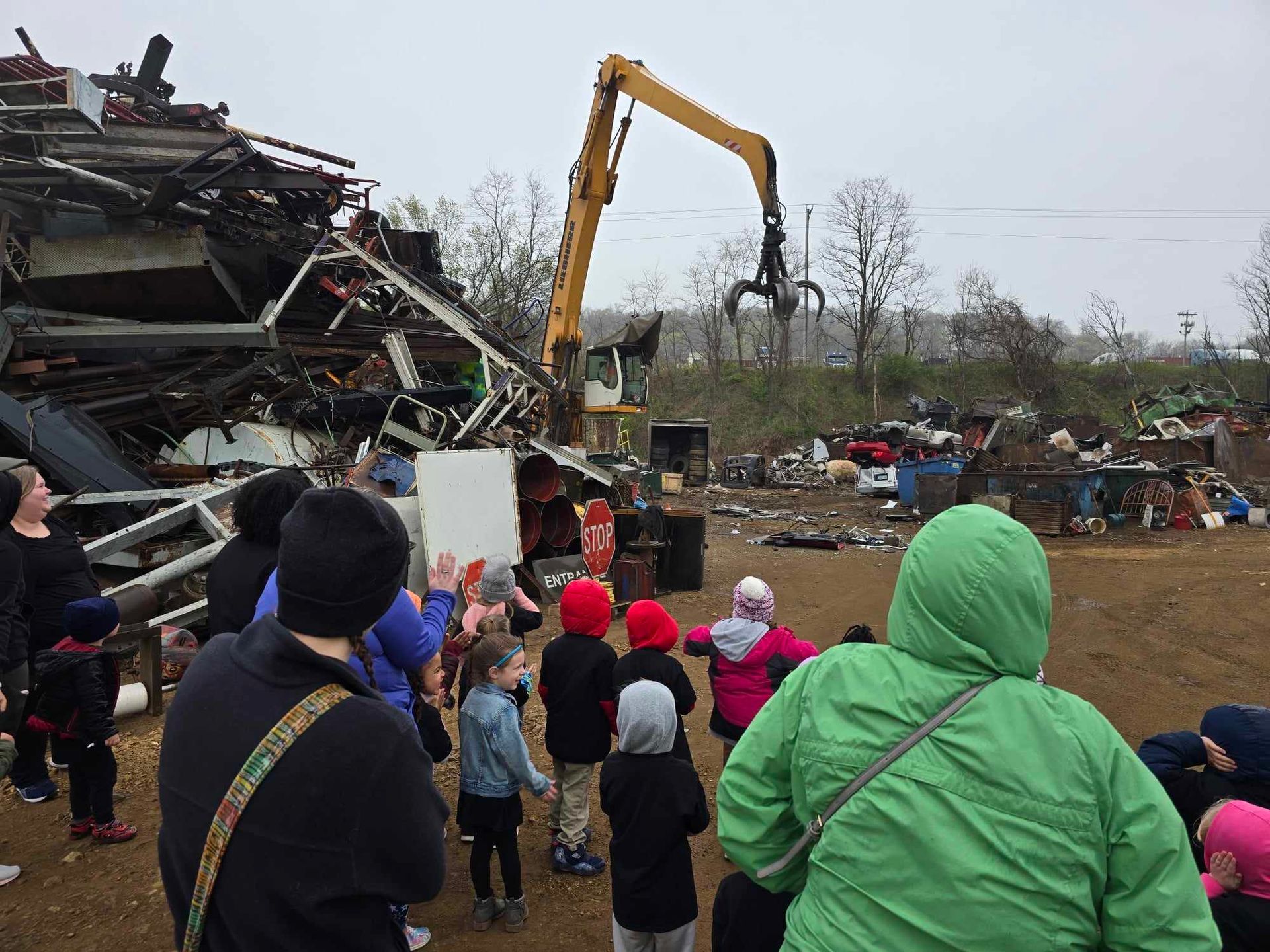 A group of people standing in a scrap yard, watching a yellow hydraulic claw machine dismantling a pile of metal debris.