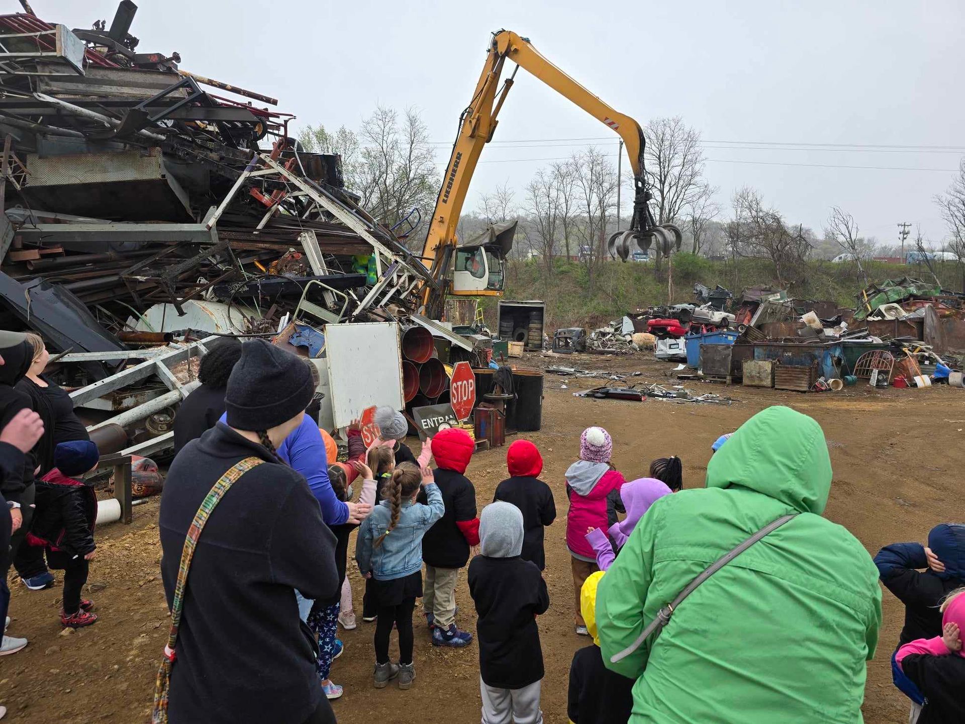 A group of people watches a large yellow excavator working at a junkyard filled with piles of debris and scrap metal.
