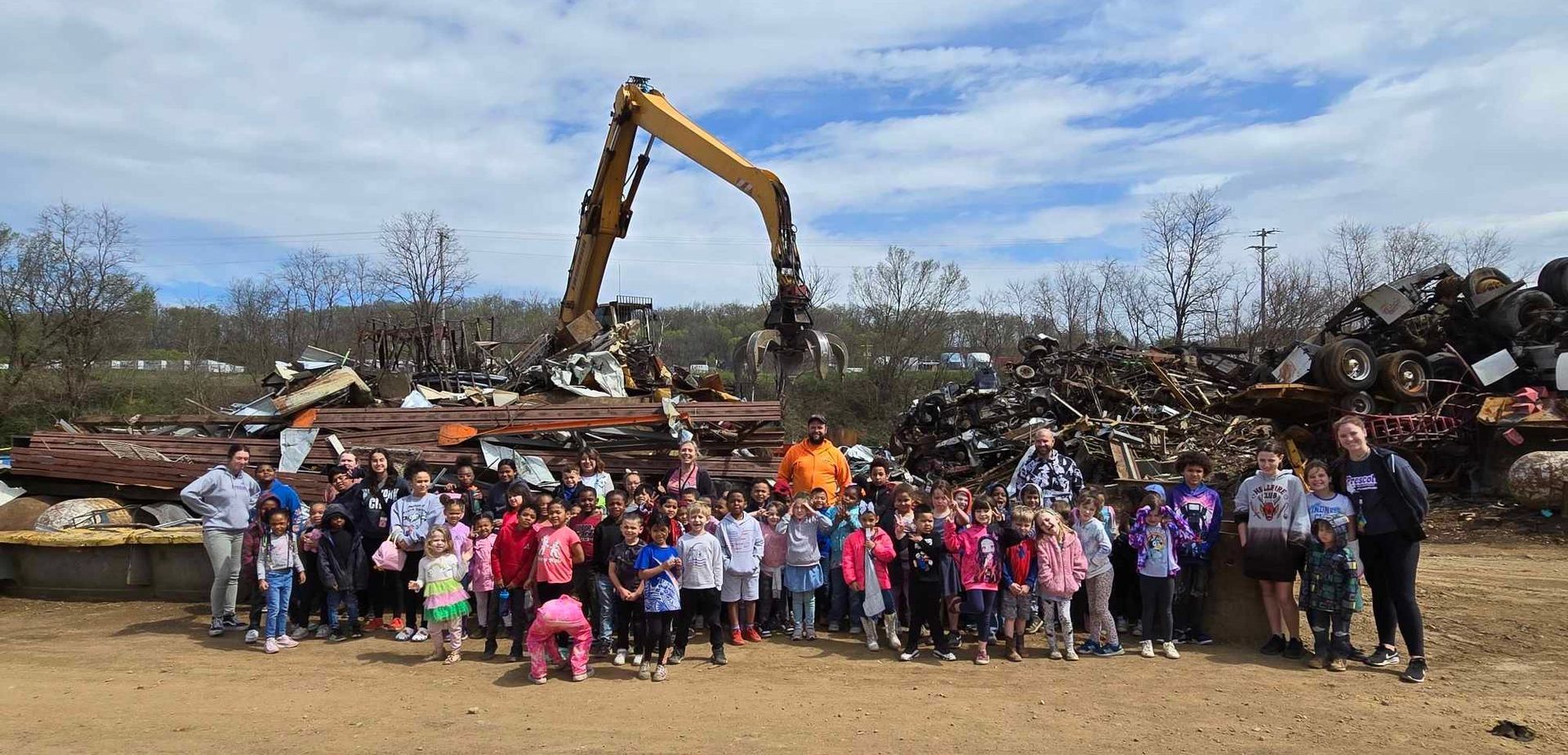 A large group poses in front of a scrap metal yard with a yellow excavator working in the background under a cloudy sky.