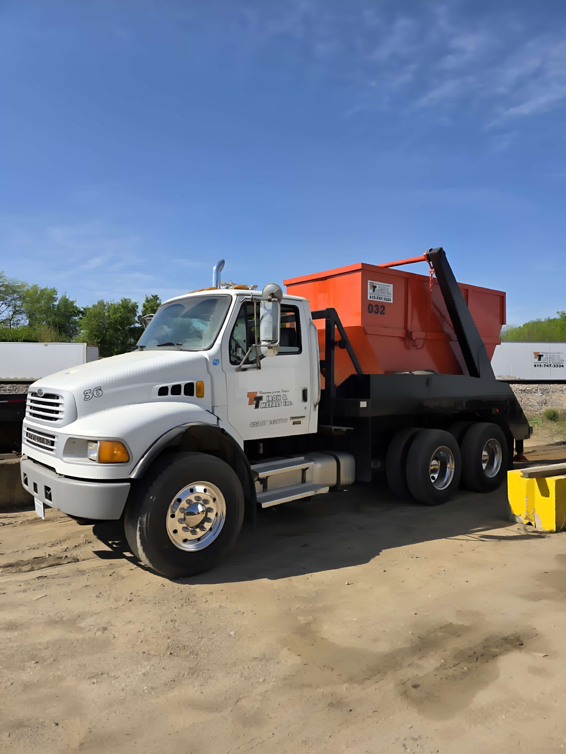 A white roll-off dump truck with an orange container parked on a dirt lot under a clear blue sky.