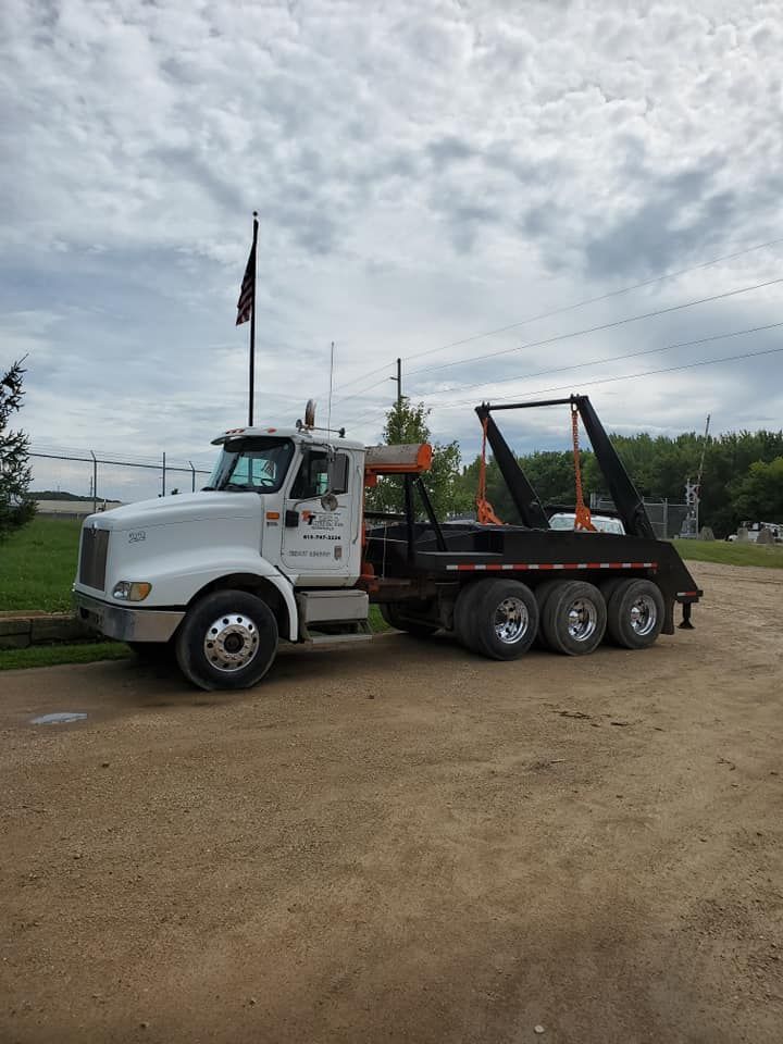 A white roll-off dump truck parked on a gravel lot under a cloudy sky with an American flag flying in the background.