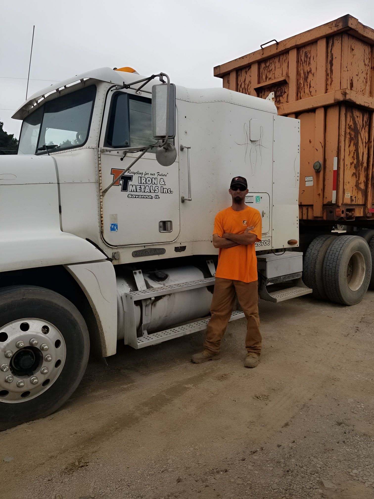 A person wearing an orange shirt and brown pants stands with arms crossed beside a white semi-truck with a rusty trailer.