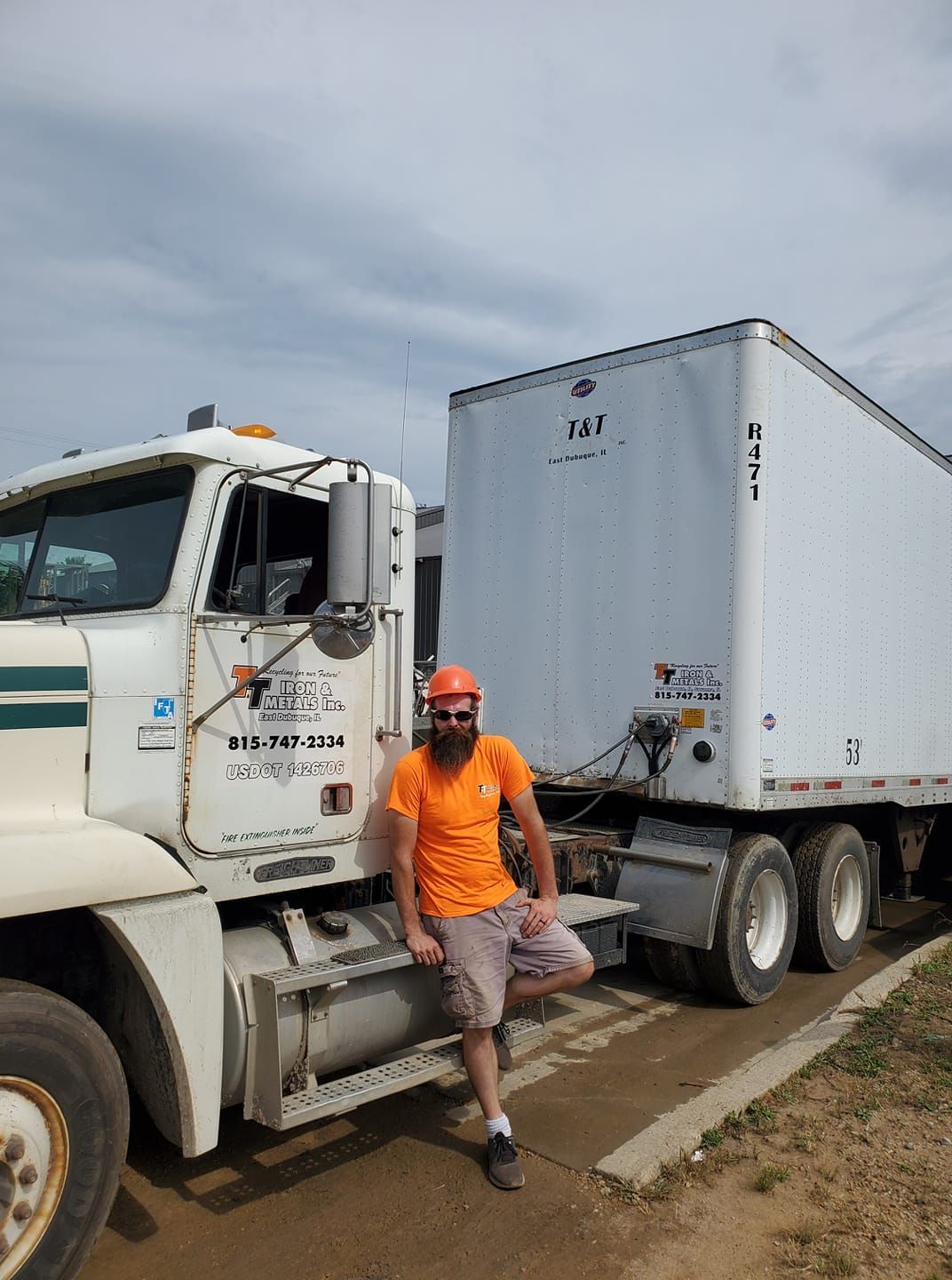 A man in an orange shirt and hard hat leans against a white semi-truck parked on a dirt lot under a cloudy sky.