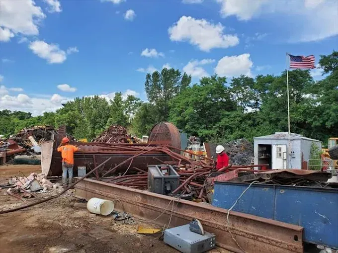 Two workers in safety gear stand among piles of rusted industrial scrap metal at an outdoor salvage yard.