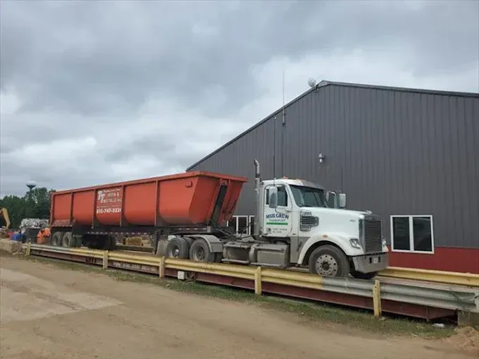 A white semi-truck pulling a long, orange dump trailer parked on a weigh scale next to a large metal industrial building.