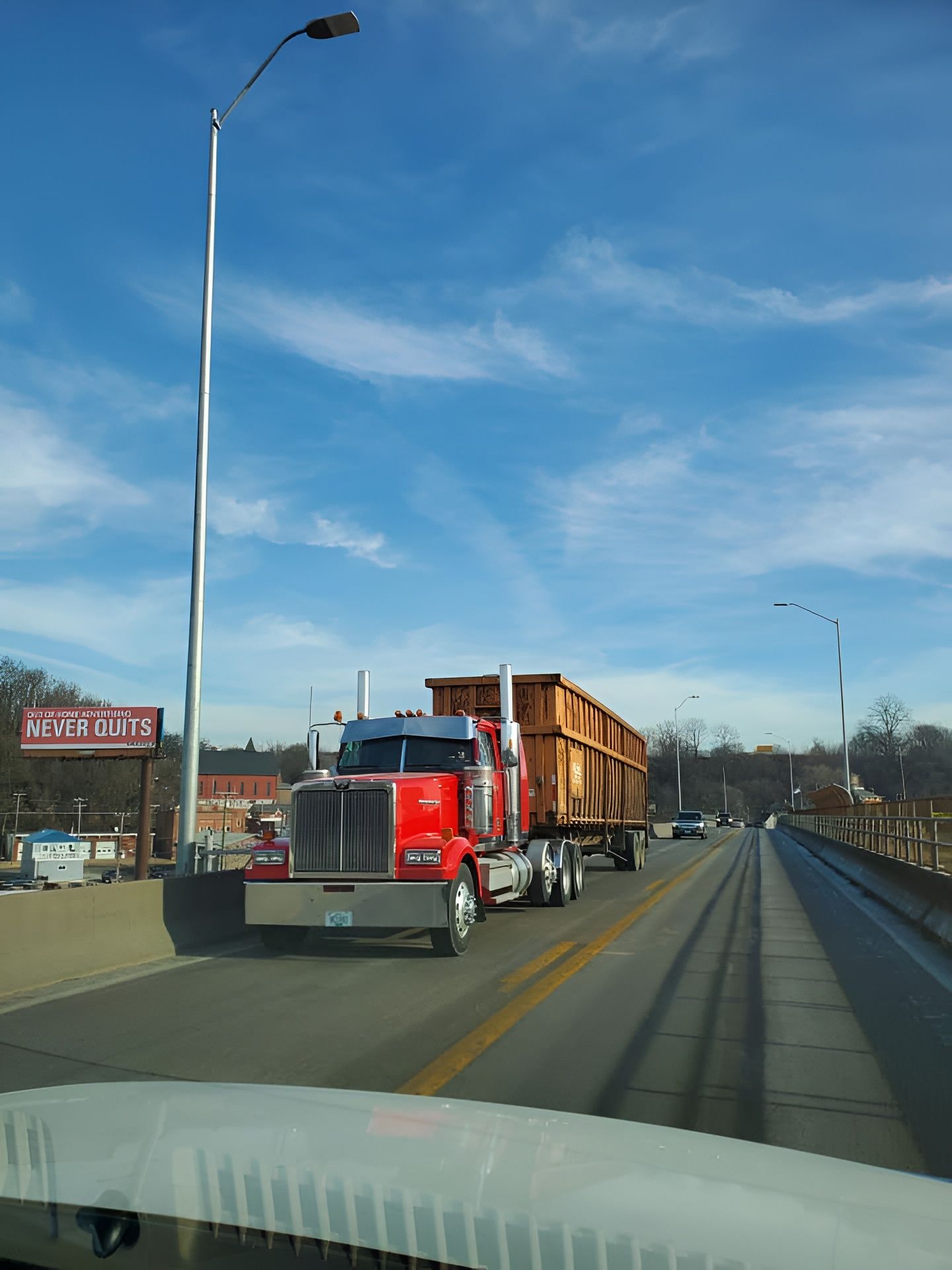 A red semi-truck carries a large wooden structure across a road under a blue, cloudy sky.