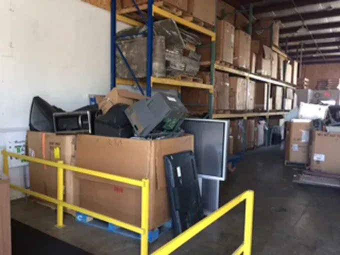 A warehouse interior featuring cardboard bins filled with discarded electronics, next to shelves stacked with boxes.