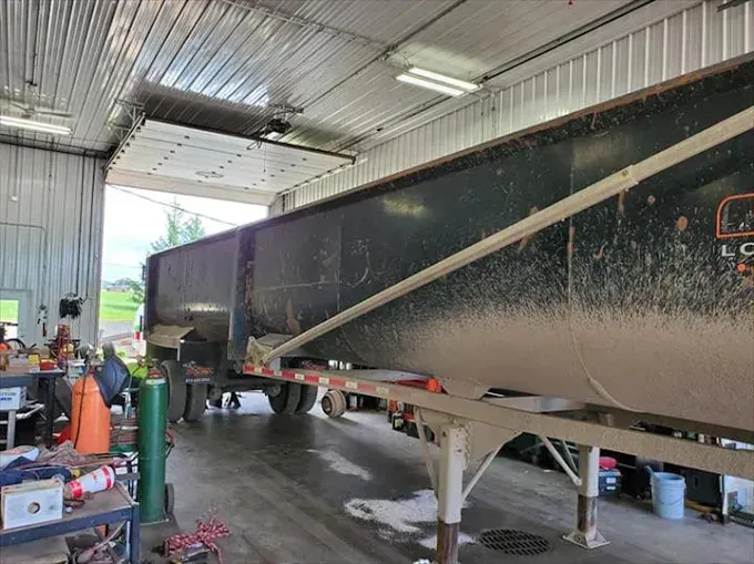 A large, black dump trailer sits inside a workshop with a raised garage door opening to a bright outdoor area.