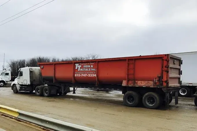 A white semi-truck pulling a long, orange metal trailer parked on a gravel lot under an overcast sky.