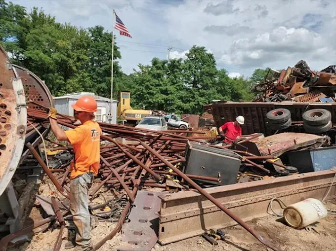 Two workers in safety gear sort through a large pile of scrap metal in an outdoor yard under an American flag.
