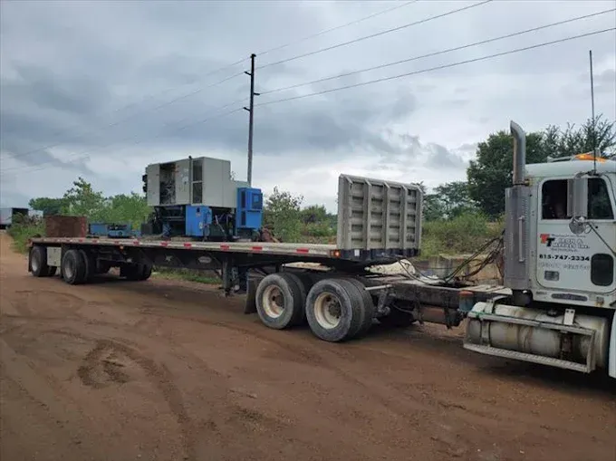A white semi-truck hauls a flatbed trailer with a blue and gray industrial generator on a dirt road under a cloudy sky.