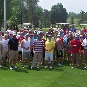 A group of people wearing casual clothing and hats stand together on a sunny outdoor golf course.