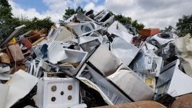A large pile of discarded white household appliances and scrap metal under a cloudy blue sky.