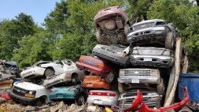 A tall, messy stack of wrecked cars and trucks in a junkyard with trees in the background.