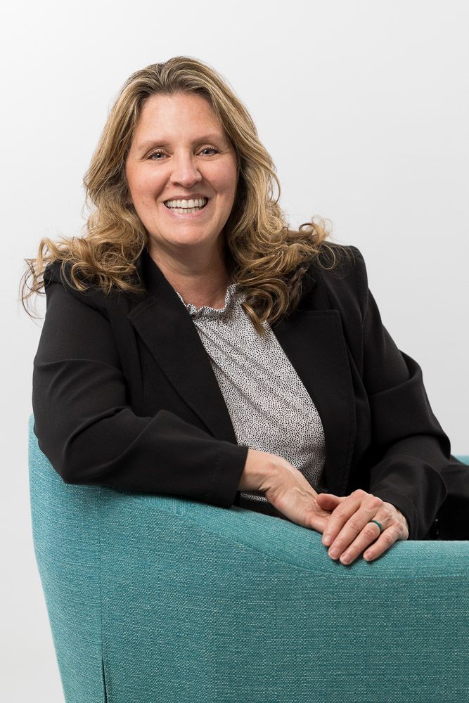 Woman with curly blonde hair wearing a black blazer sits in a teal chair, smiling.